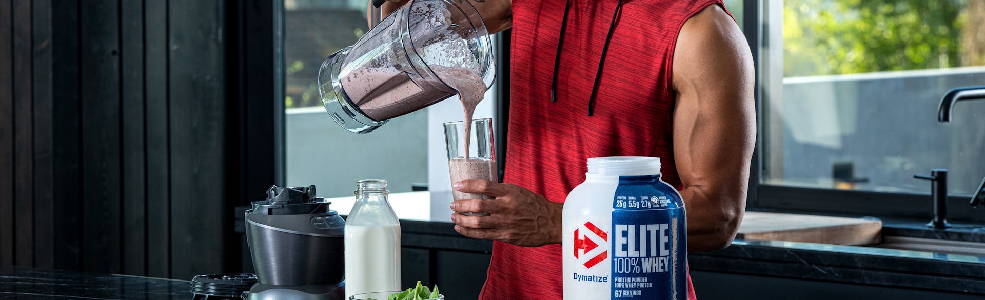 A man in a kitchen pouring a Dymatize Elite 100% Whey protein shake from a blender.