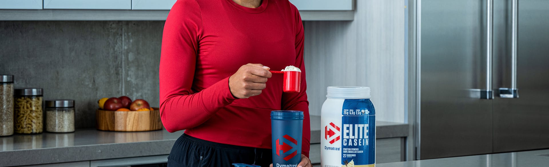 A woman in a kitchen pouring Dymatize Elite Casein powder into a shake bottle.