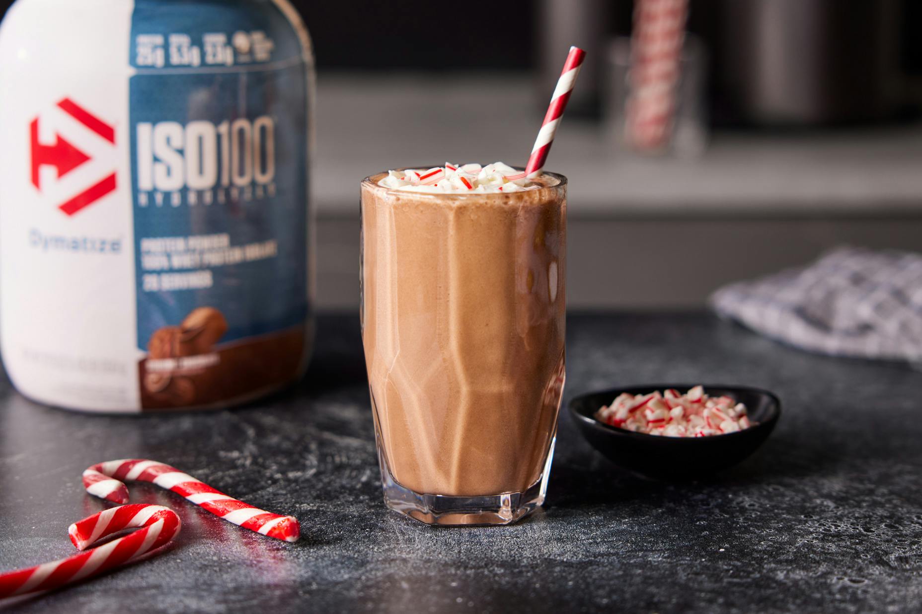 Chocolate Peppermint Protein Shake in a glass with festive straw. A tub of Dymatize and candy canes are nearby.
