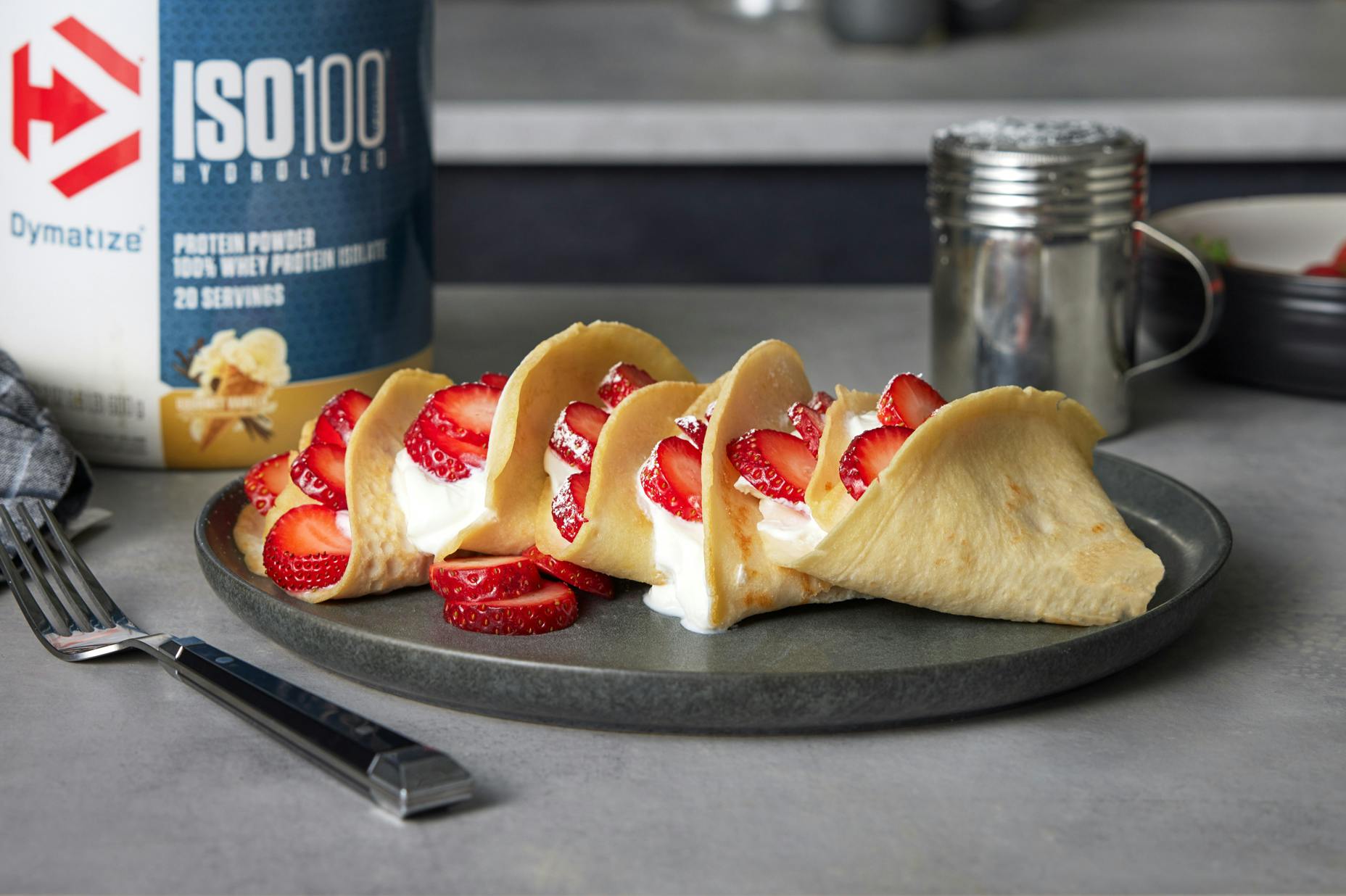 Five strawberry crepes on a plate with whipped cream. A tub of vanilla Dymatize ISO100 is on the table nearby