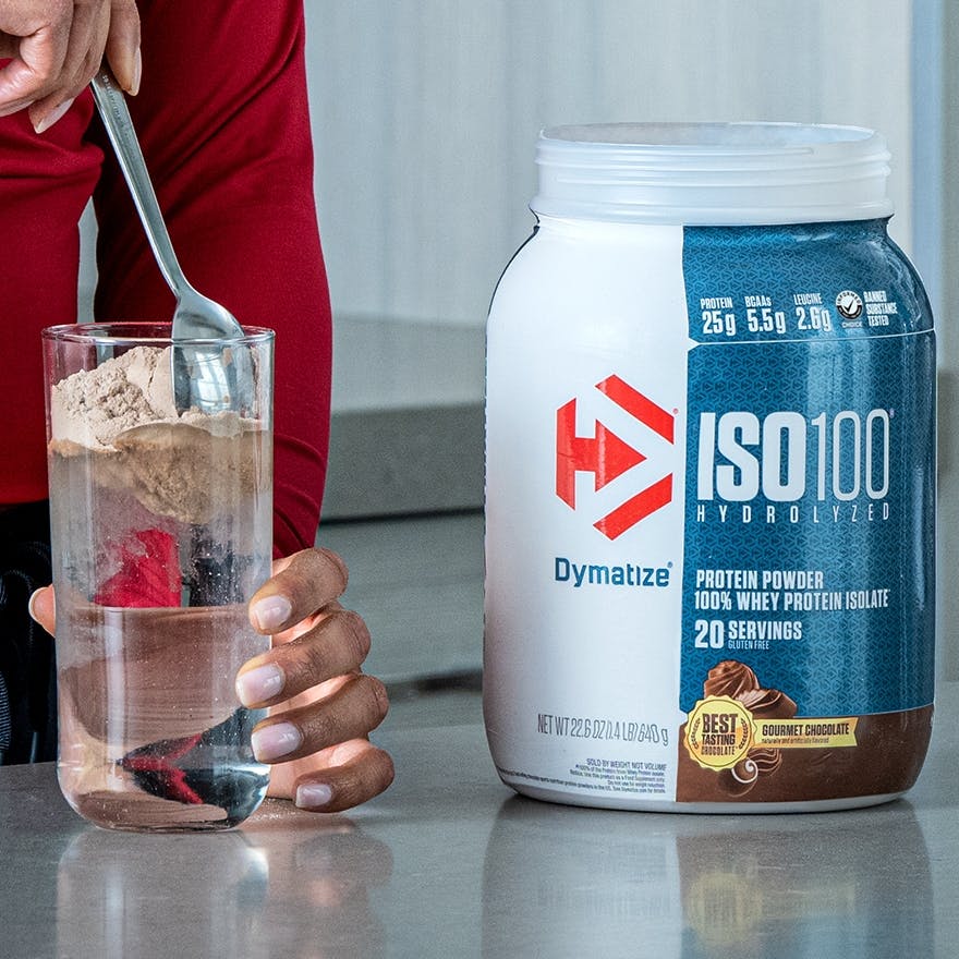 A person in a kitchen, stirring Dymatize ISO100 powder into a glass.